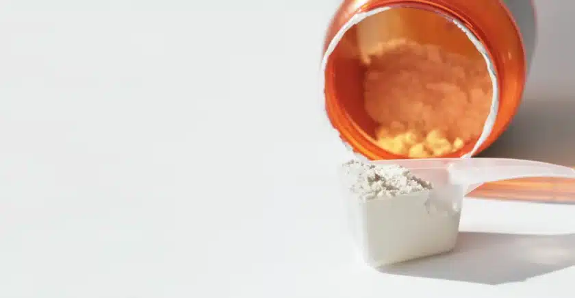 Close-up of a scoop filled with whey protein powder placed next to an open jar of protein powder on a white surface